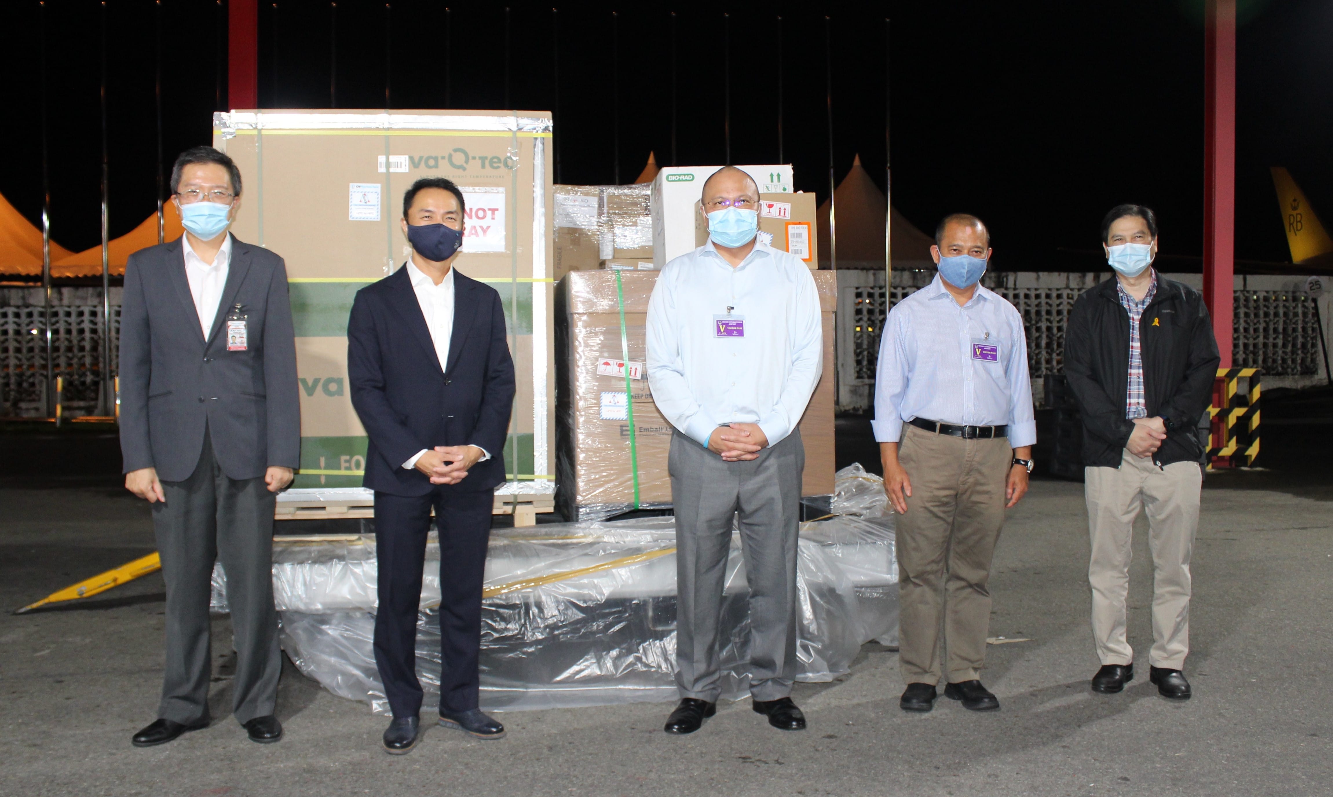 Five masked people stand before boxed cargo labeled "va-Q-tec", "BIO-RAD", and hazard symbols.